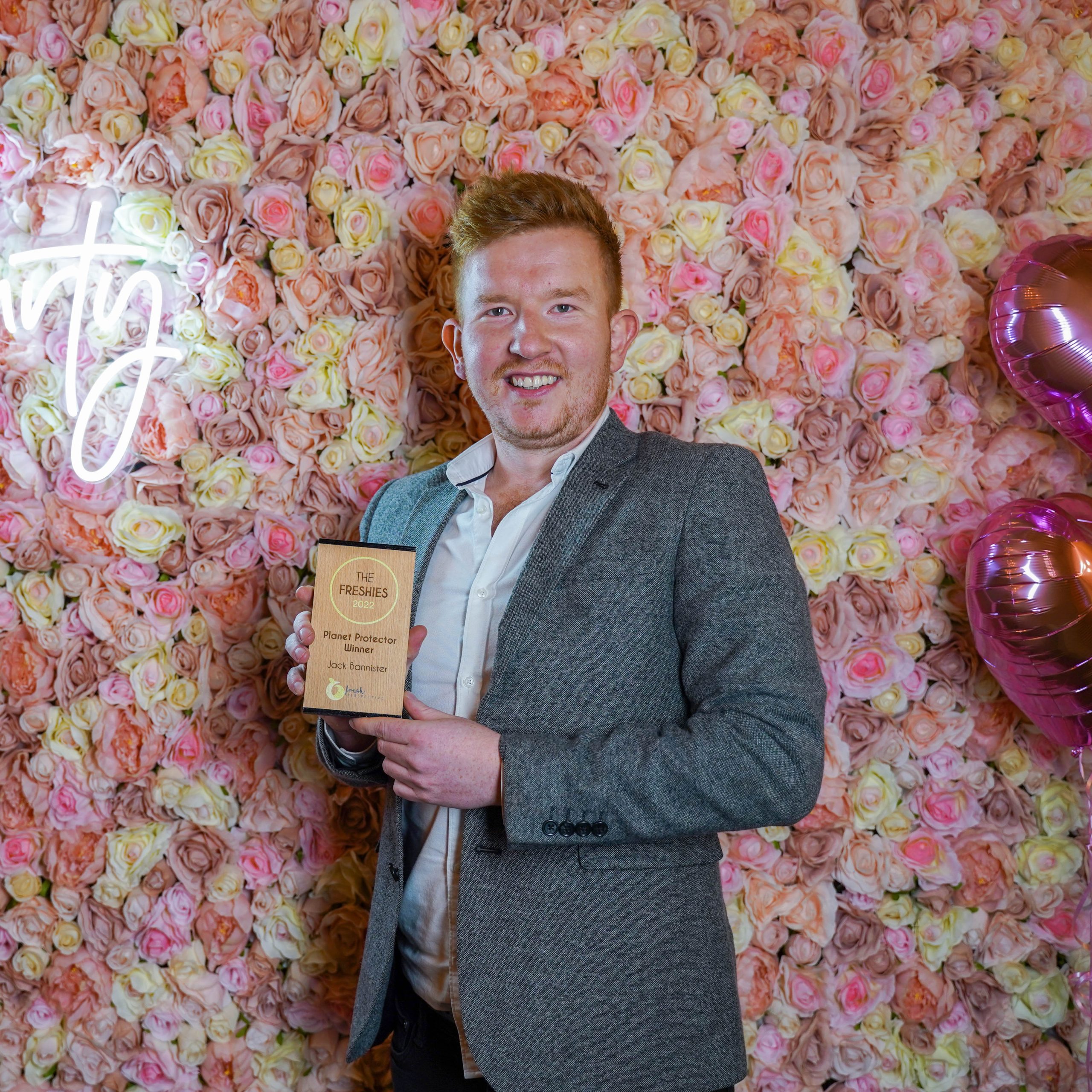 The Freshies 2022 Planet Protector winner - Jack Bannister. Stood in front of a floral wall, two pink heart balloons and a neon sign. Jack is holding his award and smiling.