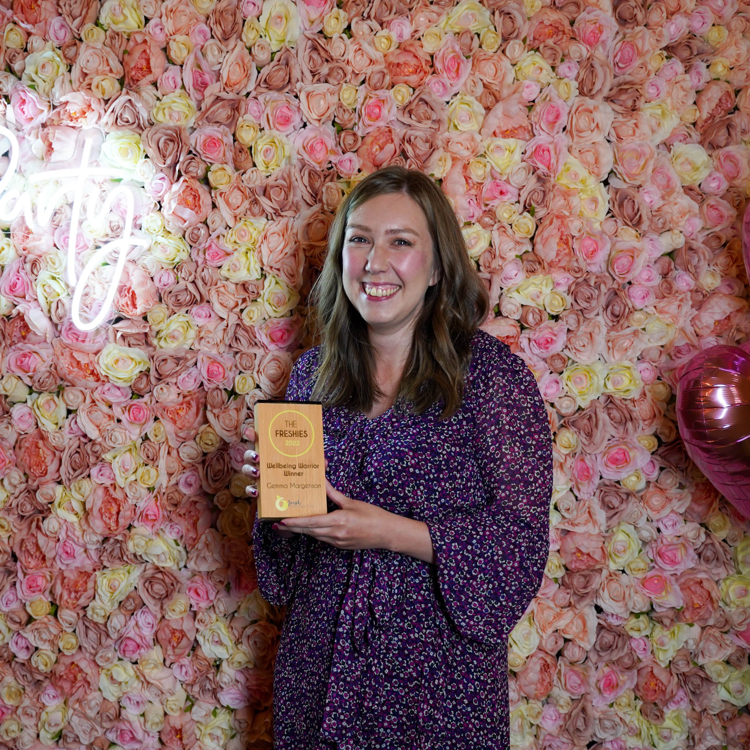 The Freshies 2022 Wellbeing Warrior winner Gemma Margerison stood in front of a pink floral wall, a pink heart balloon and a neon light sign. She is holder her award.