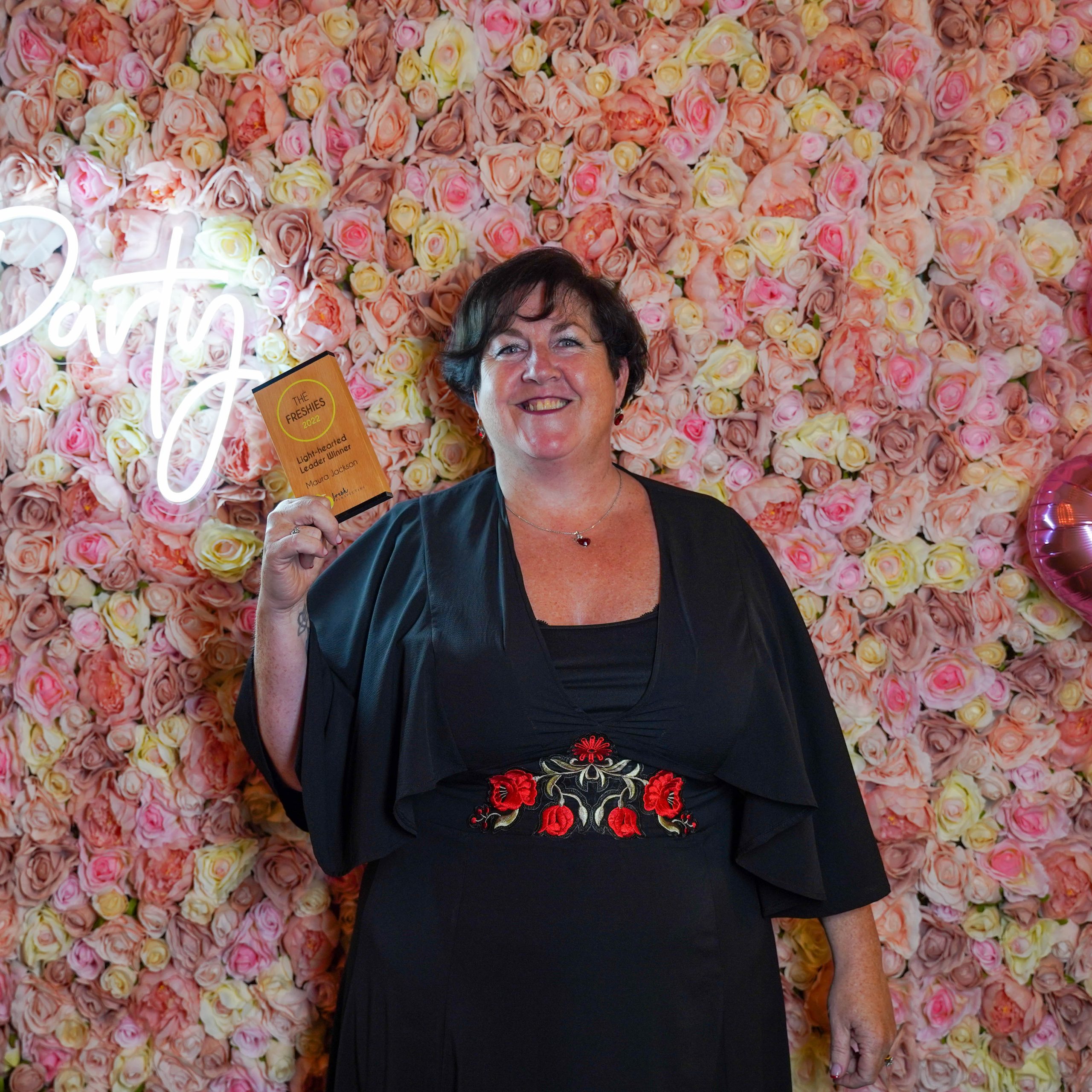 The Freshies 2022 winner for Light-hearted Leader, Maura Jackson stood in front of a pink floral wall, holding her award