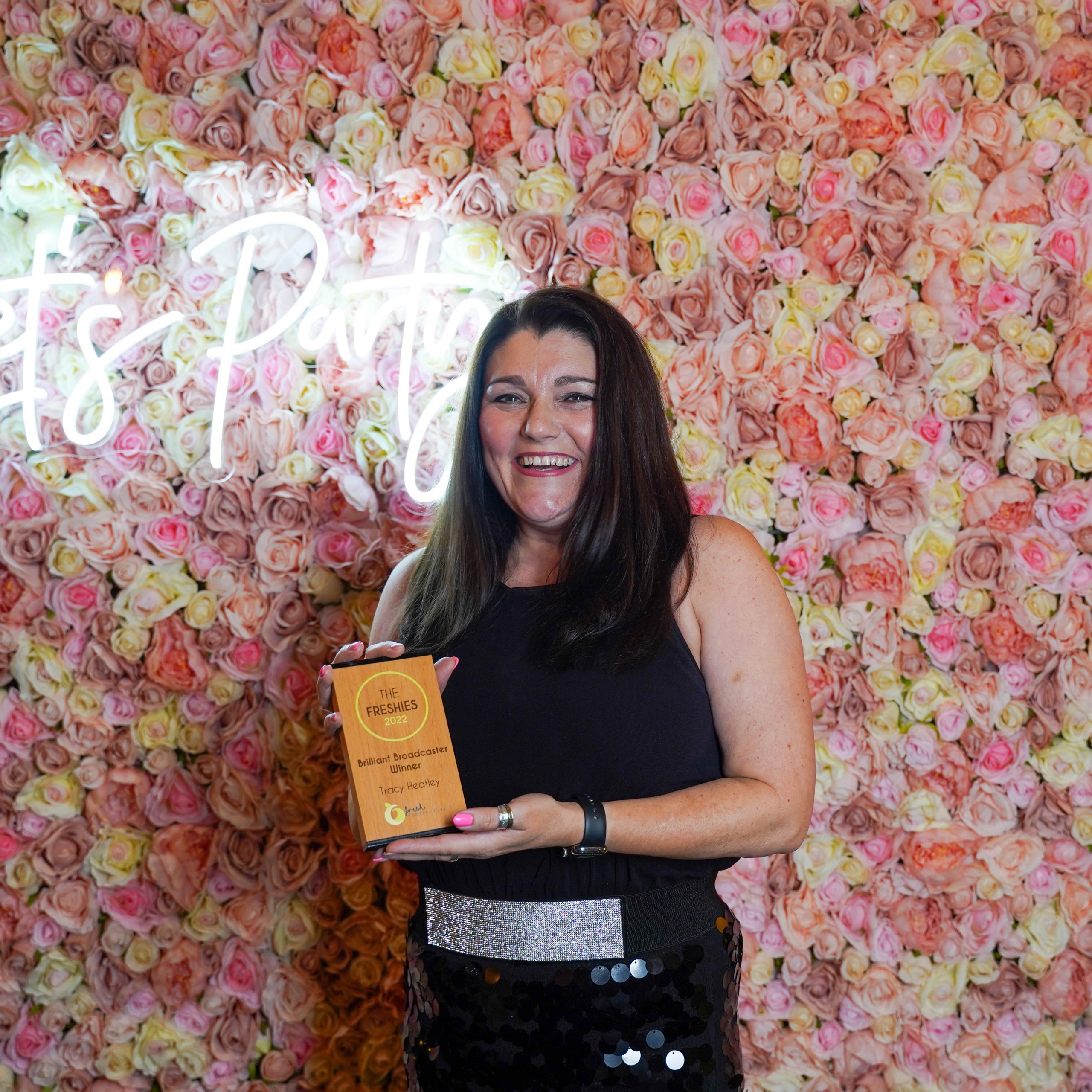 The Freshies 2022 Brilliant Broadcaster award winner Tracy Heatley, stood in front of a pink floral wall and a neon sign. She is holder her award.