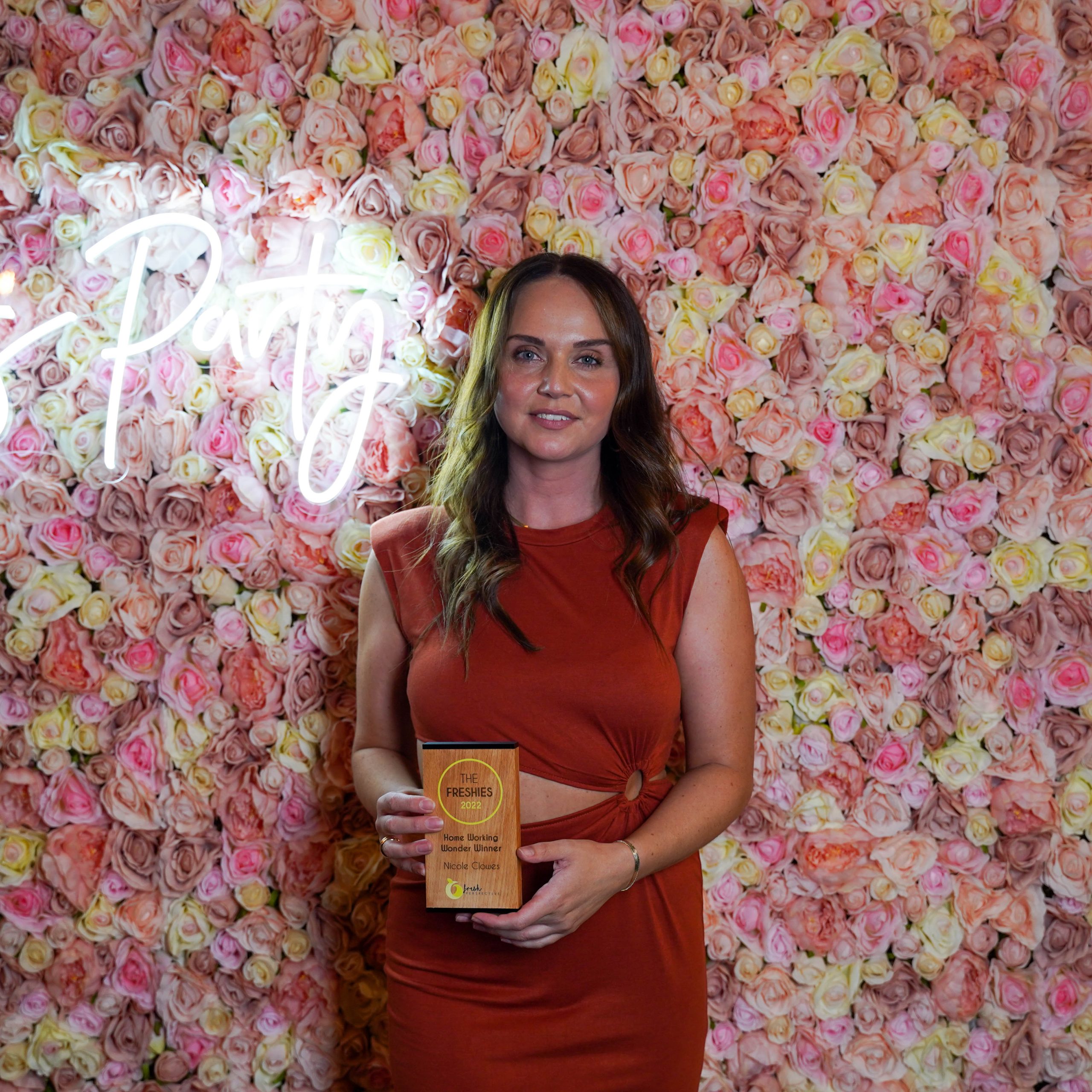 The Freshies 2022 Home Working Wonder award winner Nicole Clowes, stood in front of a pink floral wall with a neon sign. She is holding her award.