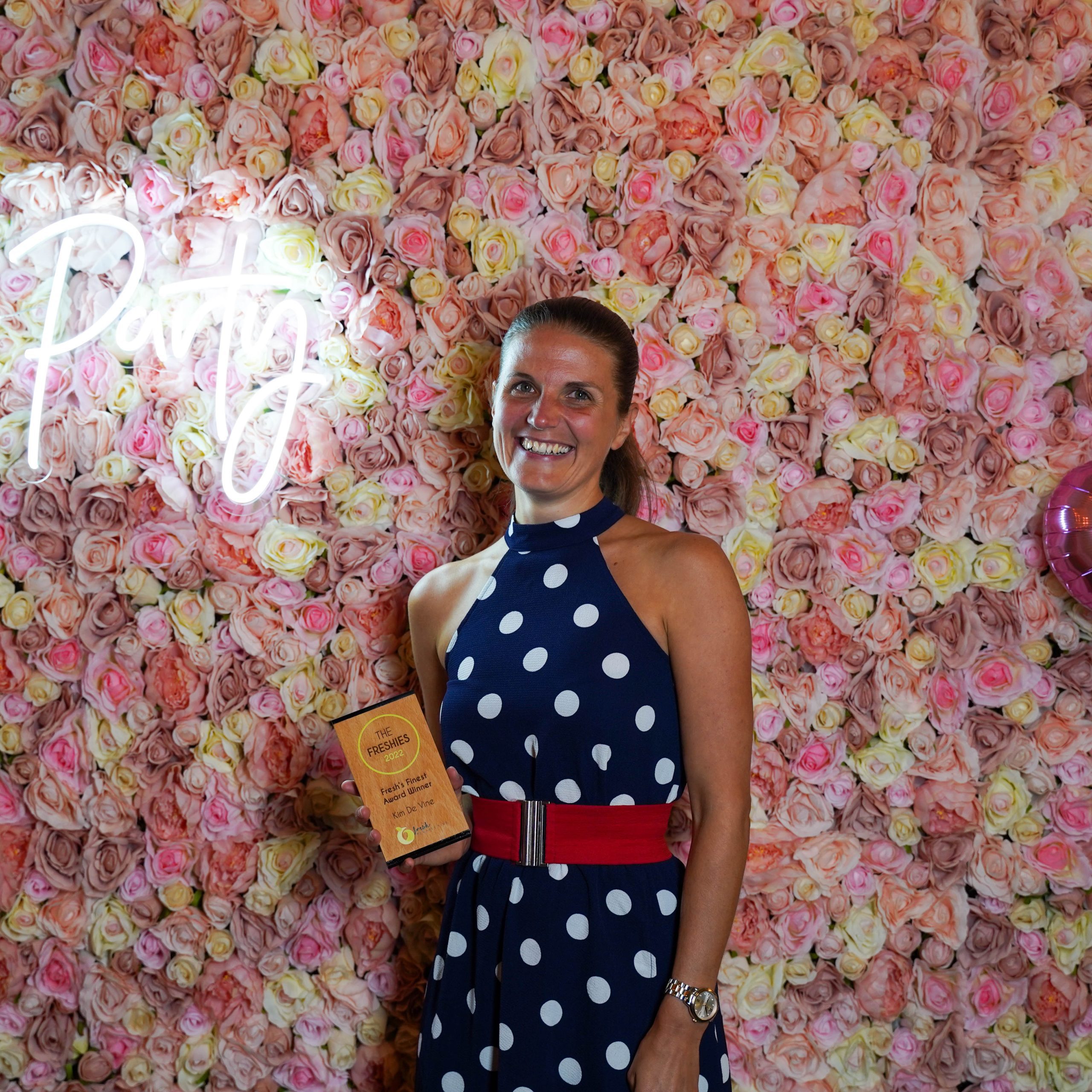 The Freshies 2022 award winner for Fresh's Finest, Kim De Vine. Kim is stood in front of a pink floral wall with a neon sign, she is stood holding her award
