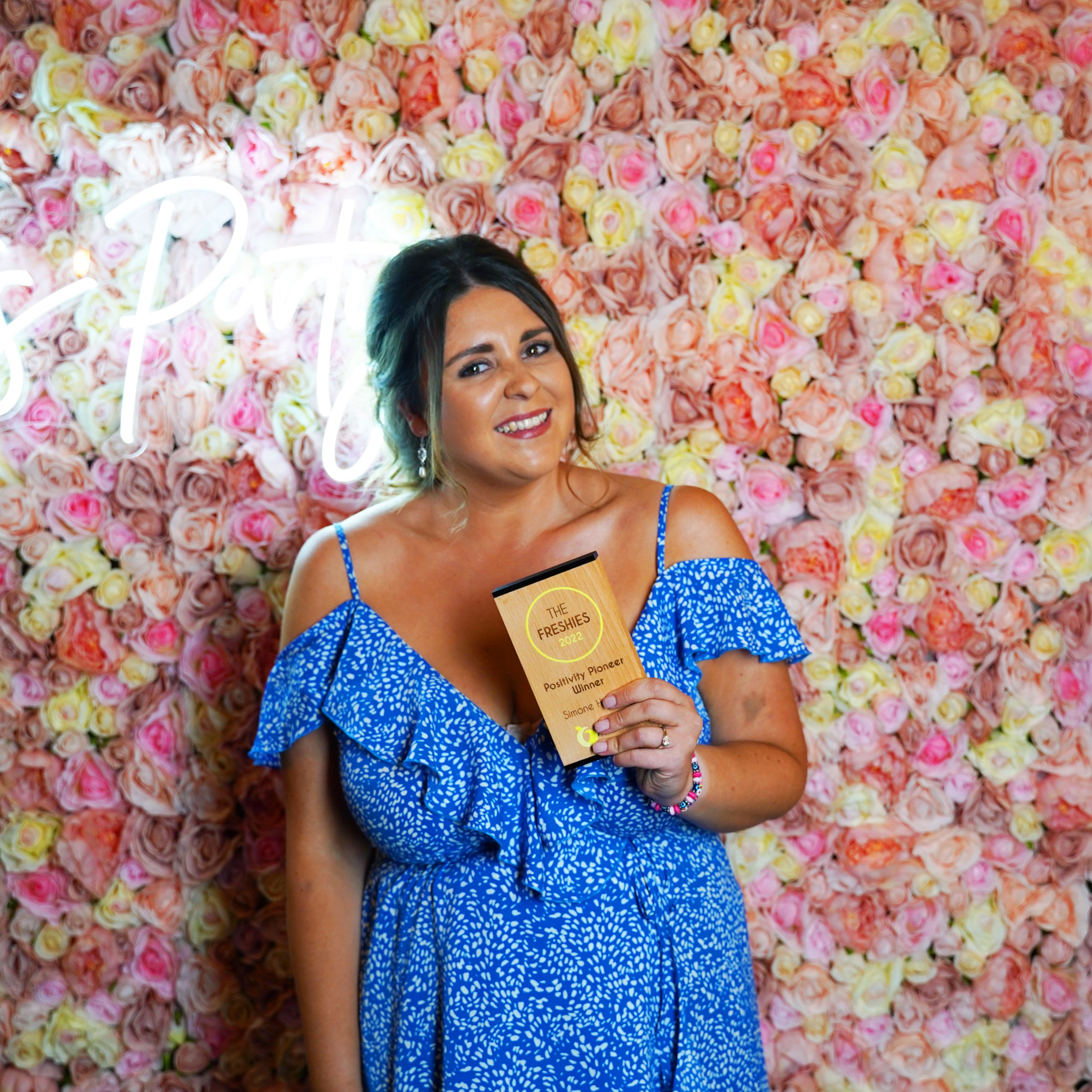 The Positivity Pioneer winner of 2022 - Simone Howarth, stood in front of a pink flower wall with two pink heart balloons and neon lighting in the back.